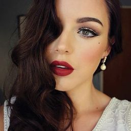 Close-up beauty portrait of a woman with dark wavy hair and vintage Hollywood glam makeup—bold red lips, winged eyeliner, beauty mark, pearl drop earring, white beaded top