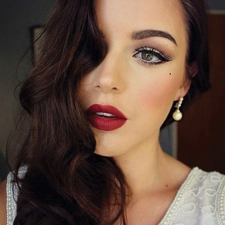 Close-up beauty portrait of a woman with dark wavy hair and vintage Hollywood glam makeup—bold red lips, winged eyeliner, beauty mark, pearl drop earring, white beaded top