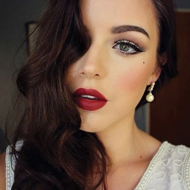 Close-up beauty portrait of a woman with dark wavy hair and vintage Hollywood glam makeup—bold red lips, winged eyeliner, beauty mark, pearl drop earring, white beaded top