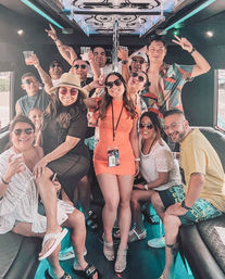 Happy group of friends partying inside a colorful party bus, posing around a center pole with drinks, sunglasses, summer outfits and neon interior lighting.