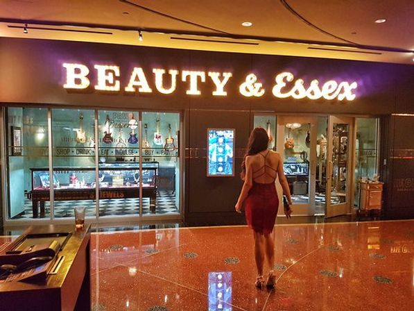 Woman in a red cocktail dress walking toward a glitzy upscale restaurant entrance with a large illuminated marquee sign, glass display windows with guitars and jewelry, and a polished terrazzo casino hallway.