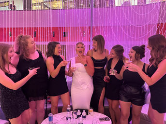 Lively bachelorette party: women in black dresses cheer as a woman in a white dress shows off a ring, holding cocktails in a pink-lit lounge with beaded curtain backdrop.
