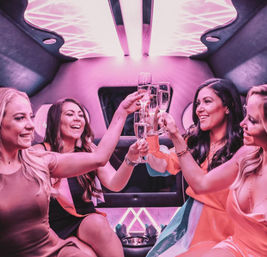 Four women laughing and toasting champagne inside a pink LED-lit limousine interior, celebrating a bachelorette party / girls' night out.