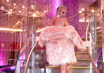 Back-facing figure in a fluffy pink faux-fur coat ascending a glass spiral staircase beneath sparkling chandeliers and magenta lighting.