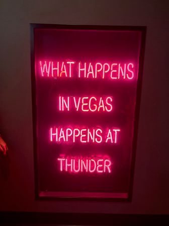 Vibrant pink neon sign reading WHAT HAPPENS IN VEGAS HAPPENS AT THUNDER, framed on a dark wall — neon Las Vegas wall art