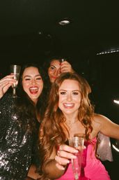 Three smiling women in party dresses raising champagne flutes inside a limo at night, celebrating a girls' night out or bachelorette party