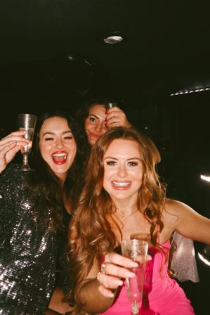 Three smiling women in party dresses raising champagne flutes inside a limo at night, celebrating a girls' night out or bachelorette party