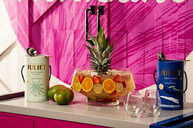 Tropical party drink station on a white countertop with a large glass punch bowl topped with a pineapple and filled with sliced oranges, limes and strawberries, two green mangoes, two packaged wine canisters and two stemless glasses against a vibrant pink backdrop.