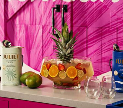 Tropical party drink station on a white countertop with a large glass punch bowl topped with a pineapple and filled with sliced oranges, limes and strawberries, two green mangoes, two packaged wine canisters and two stemless glasses against a vibrant pink backdrop.