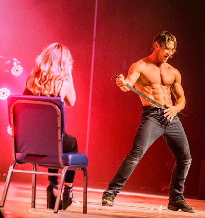 Shirtless male dancer performing a theatrical striptease on a red-lit stage, pulling a belt while a seated woman in heels watches from a chair — live cabaret/club performance.