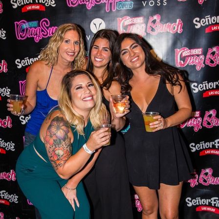 Four smiling women holding plastic cups and posing at a Las Vegas drag-brunch photo wall, friends dressed for a night out — one woman in green shows a colorful floral arm tattoo.