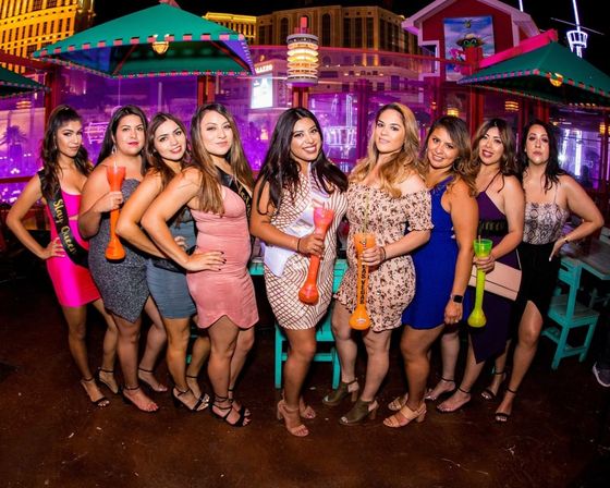 Las Vegas nightlife bachelorette party: group of women in cocktail dresses holding colorful yard‑long cocktails, posing at a neon‑lit rooftop bar with casino skyline in the background.