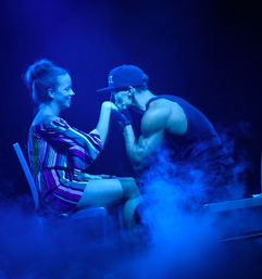 Male performer in a cap kisses a seated woman's hand during an intimate live stage show with blue lighting and fog