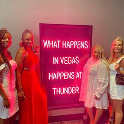 Four women in party dresses and tiaras pose and smile beside a bright pink neon sign with a Vegas slogan, capturing a lively Las Vegas nightlife moment.