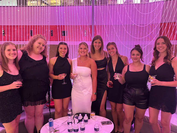 Eight women in evening dresses, a bride-to-be in white wearing a sash, holding cocktails and smiling in front of a pink beaded curtain backdrop at a bachelorette party lounge.