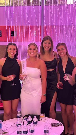 Eight women in evening dresses, a bride-to-be in white wearing a sash, holding cocktails and smiling in front of a pink beaded curtain backdrop at a bachelorette party lounge.