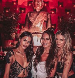 Three women in stylish evening outfits smiling and posing in a red-lit lounge or restaurant with a large seated Buddha statue and candle-lit niches behind them, nightlife dining vibe.