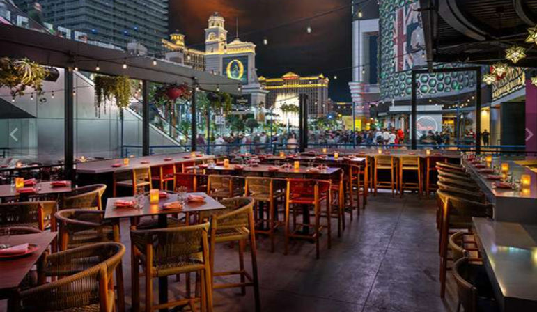 Outdoor rooftop dining terrace at night with wooden tables, wicker chairs, candlelit place settings and string lights overlooking a bustling neon-lit city street and illuminated hotel towers — outdoor dining, city nightlife.