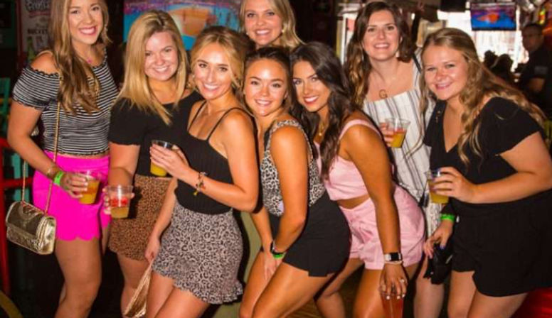 Group of eight smiling women posing with drinks during a lively night out at a crowded city bar, colorful lights and summer party outfits