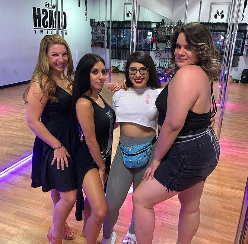 Four women posing together in a pole-dance studio group photo — mirrored walls, chrome poles and wooden floors, wearing casual workout and night-out outfits.
