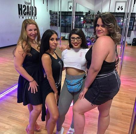 Four women posing together in a pole-dance studio group photo — mirrored walls, chrome poles and wooden floors, wearing casual workout and night-out outfits.