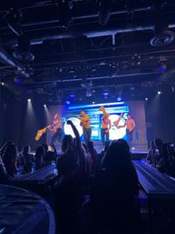 Shirtless male dancers perform a high-energy nightclub cabaret routine under blue stage lights and an LED boombox backdrop while a silhouetted audience films.