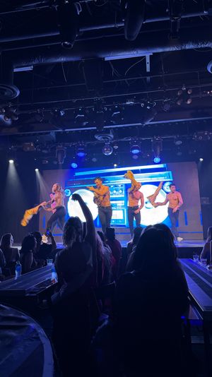 Shirtless male dancers perform a high-energy nightclub cabaret routine under blue stage lights and an LED boombox backdrop while a silhouetted audience films.