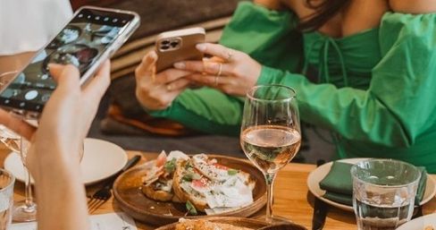 Cafe brunch scene: two diners using smartphones to photograph open-faced toasts topped with cheese and greens, a glass of wine and water on a wooden table — Instagram-ready meal.
