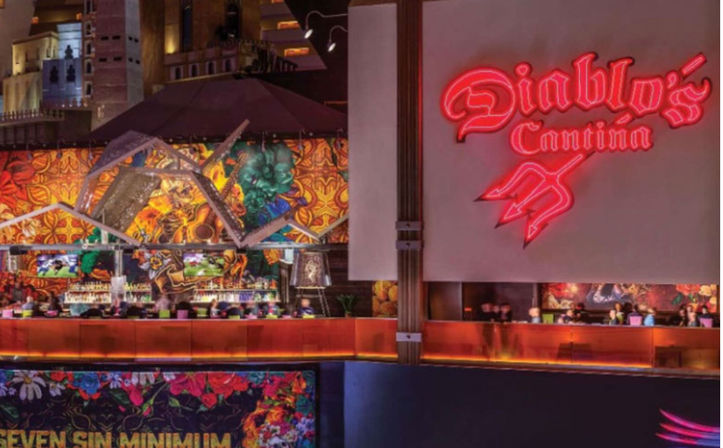 Vibrant Mexican-style cantina interior with a large neon red script sign above a long illuminated bar, colorful floral murals and decorative accents, and patrons seated amid warm, festive lighting.