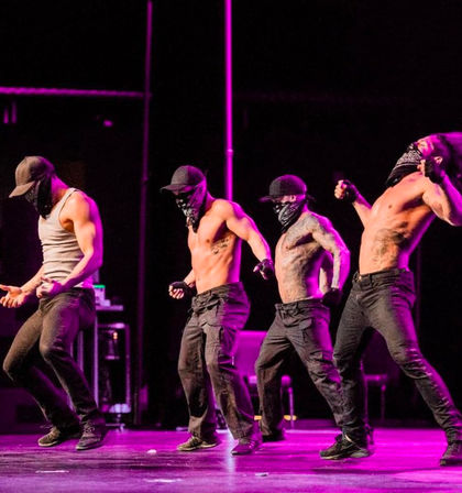 Four masked male dancers in caps and bandanas performing a high-energy choreographed routine on a purple-lit stage, wearing tank tops or shirtless with cargo pants.