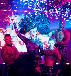 Drag queen in a sparkly black lace gown and giant feather headdress performs on a neon-lit nightclub stage with shirtless backup dancers as colorful confetti rains down