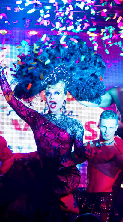 Drag queen in a sparkly black lace gown and giant feather headdress performs on a neon-lit nightclub stage with shirtless backup dancers as colorful confetti rains down