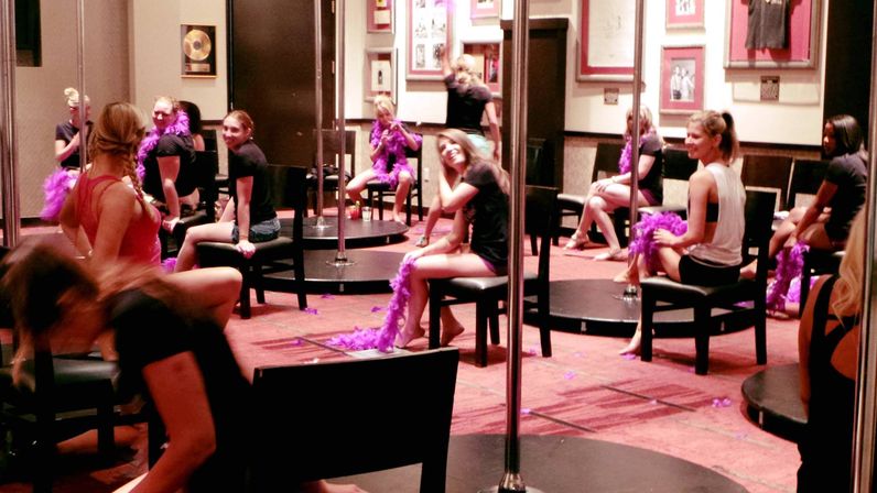 Group of women in a playful indoor pole-dance class using chairs and purple feather boas in a dance studio
