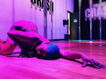 Dancer with short blue hair stretched across a wood floor, reaching forward under magenta-purple lighting beside chrome poles in an indoor pole-dance studio.
