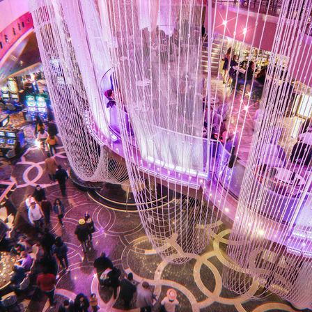 Vibrant purple-lit casino atrium seen from above, beaded chandelier draping over a busy multi-level gaming floor with slot machines, patterned marble walkways, and an elevated bar lounge.