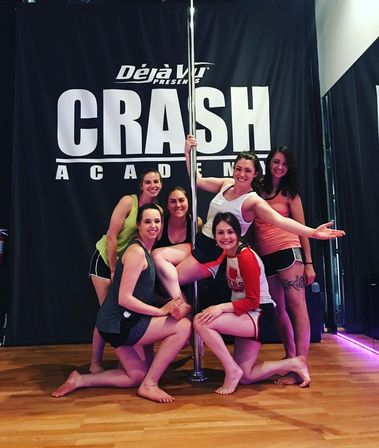 Six women smiling and posing around a chrome pole in a wood‑floored pole dance studio, wearing athletic shorts and tank tops — group pole dance fitness class.