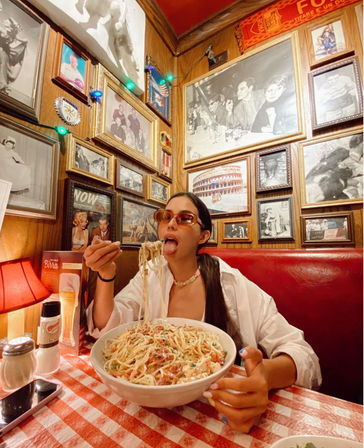 Playful diner about to taste a giant bowl of spaghetti in a cozy Italian restaurant booth with a red checkered tablecloth and wood-paneled walls covered in vintage framed photos and memorabilia.