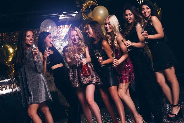 Seven women in sparkly cocktail dresses laughing and toasting with champagne during a girls' night out in a dimly lit club with gold balloons and confetti.