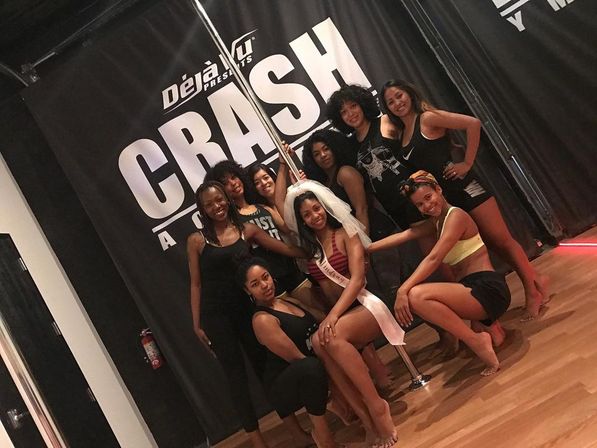 Group of smiling women in an indoor pole-dance studio posing around a chrome pole, bride-to-be wearing a veil and sash at center with wood floor and black backdrop.