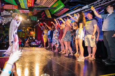 Drag performer on a nightclub stage leading a lively toast as a line of patrons raise shot glasses beneath colorful hanging signs.