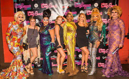 Group photo of drag performers and fans in glittering gowns, bold makeup and high heels posing on a red‑carpet style backdrop at a lively drag show event