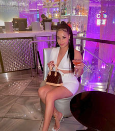 Woman in a white two-piece dress and strappy heels seated in an upscale lounge with purple ambient lighting, holding a cocktail and a brown monogram designer handbag, modern glass bar and mirrored décor in the background.
