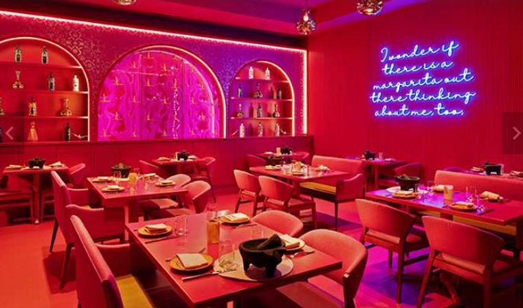 Vibrant pink neon-lit restaurant interior with arched backlit bar shelves, tables set for dining, plush chairs and a blue neon wall sign reading "I wonder if there is a margarita out there thinking about me, too."