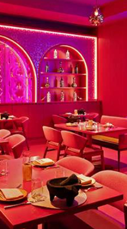 Vibrant pink neon-lit restaurant interior with arched backlit bar shelves, tables set for dining, plush chairs and a blue neon wall sign reading "I wonder if there is a margarita out there thinking about me, too."