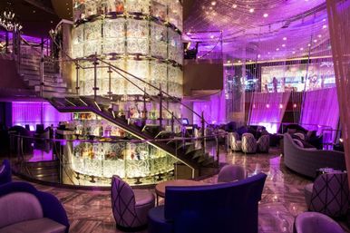 Glitzy purple-lit luxury hotel lounge and bar interior with illuminated central cylindrical display, spiral glass staircase, cascading crystal bead curtains, plush curved seating and glossy marble floor.