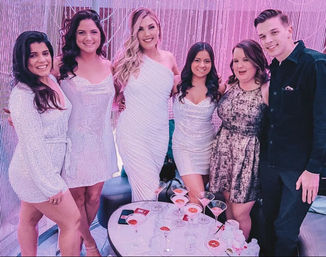 Six friends dressed up pose under pink lights at a cocktail lounge with a beaded backdrop, sparkly white dresses and one man in a dark shirt gathered around a table of martini glasses — lively night-out group photo.