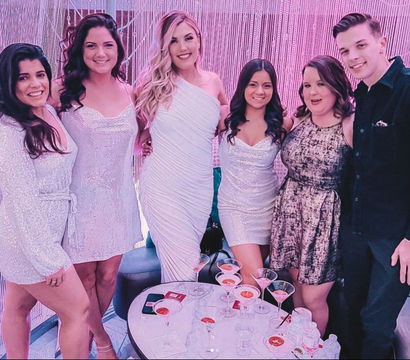 Six friends in sparkly party attire pose in front of a pink‑lit sequin backdrop at a cocktail lounge, smiling around a table of martini glasses and bottled water.