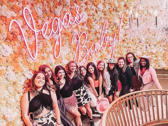 Smiling group of women posing on a curved staircase in front of a peach-and-white floral wall with a neon "Vegas Baby!" sign, wearing festive outfits, cow-print skirts and pink cowboy hats for a girls' night out.