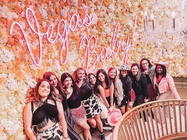 Smiling group of women posing on a curved staircase in front of a peach-and-white floral wall with a neon "Vegas Baby!" sign, wearing festive outfits, cow-print skirts and pink cowboy hats for a girls' night out.