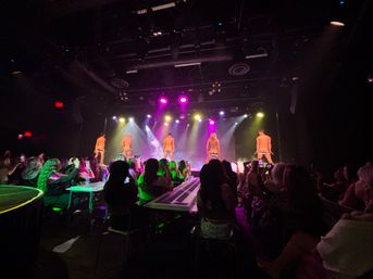 Packed nightclub cabaret: five male dancers on stage facing away, dropping underwear under colorful spotlights and haze while a seated audience watches and records on phones.